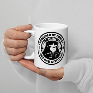 May include: White ceramic mug with a black and white illustration of a bear drinking coffee. The mug has the text "Powered by Coffee and Bad Attitudes" around the bear.