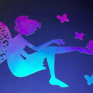 Pode incluir: Uma silhueta de fada iridescente azul e roxa com asas e um design de borboleta. A fada está sentada com uma perna estendida e segurando uma borboleta na mão. Há quatro borboletas menores no fundo.
