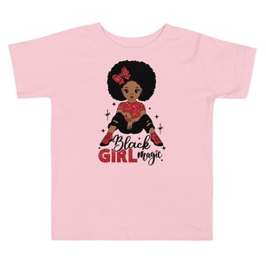 May include: Pink toddler t-shirt with a black girl cartoon illustration wearing a red bow and red shoes. The text "Black Girl Magic" is written in red glitter letters.