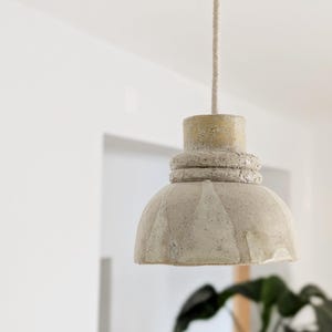 Puede incluir: Una lámpara colgante con una pantalla de cerámica texturizada en color blanco roto, con un perfil en forma de campana y un cuello grueso y estriado en la parte superior. La lámpara cuelga de un cable de tela claro que sube desde el centro.