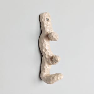 Peut inclure: Un porte-manteau mural vertical avec une surface texturée et alvéolée de couleur crème pâle. L'objet présente trois crochets saillants disposés en colonne, avec une tête de vis métallique visible en haut pour la fixation au mur.