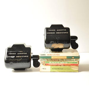 May include: Two black electrical switches with the text "ΓΕΝΙΚΟΣ ΔΙΑΚΟΠΤΗΣ ΚΙΝΗΣΕΩΣ ΑΝΕΛΚΥΣΤΗΡΟΣ" and "40144500V" on them. The switches are sitting on top of a stack of paperback books.