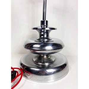 Op de afbeelding: Een chromen tafellamp met een gelaagde basis en een enkele gloeilamp. De lamp heeft een rood snoer.