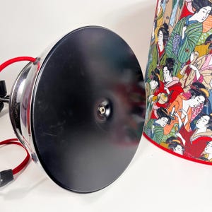 Op de afbeelding: Een zwarte metalen lampvoet met een rood snoer en een ronde, zwarte metalen kap. De lampenkap heeft een kleurrijk ontwerp met Japanse vrouwen in traditionele kleding.