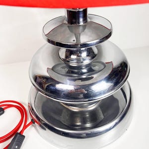 Op de afbeelding: Een chromen tafellamp met een gelaagde basis en een rode lampenkap met een bloemenprint.