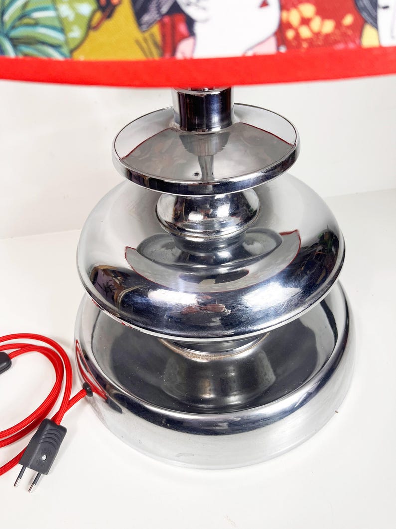 Op de afbeelding: Een chromen tafellamp met een gelaagde basis en een rode lampenkap met een bloemenprint.