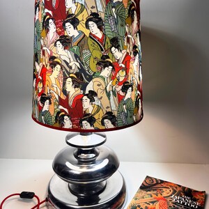 Op de afbeelding: Een zilveren tafellamp met een rood-witte lampenkap met een herhalend patroon van Japanse vrouwen. De lampenkap is cilindrisch en heeft een rode rand. De lampvoet bestaat uit drie gestapelde zilveren schijven. Op de voorgrond is een boek te zien met de titel "The Japanese Tattoo".