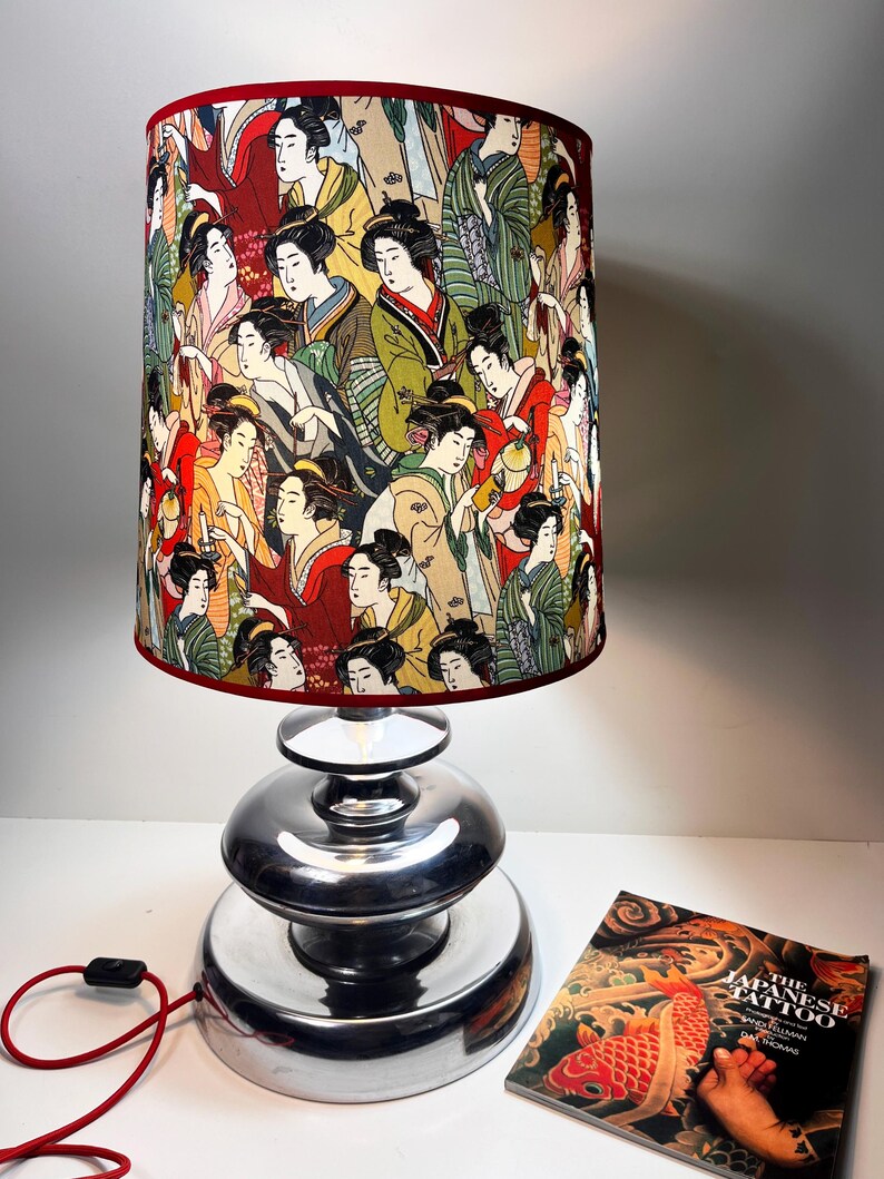 Op de afbeelding: Een zilveren tafellamp met een rood-witte lampenkap met een herhalend patroon van Japanse vrouwen. De lampenkap is cilindrisch en heeft een rode rand. De lampvoet bestaat uit drie gestapelde zilveren schijven. Op de voorgrond is een boek te zien met de titel "The Japanese Tattoo".