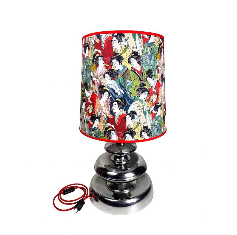 Op de afbeelding: Een zilveren tafellamp met een gestapelde basis en een rood-witte lampenkap met een patroon van Japanse vrouwen in traditionele kleding.