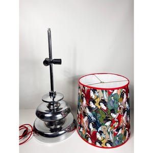 Op de afbeelding: Een chromen tafellamp met een gelaagde basis en een rood-witte lampenkap met een ontwerp van vrouwen in traditionele Japanse kleding.