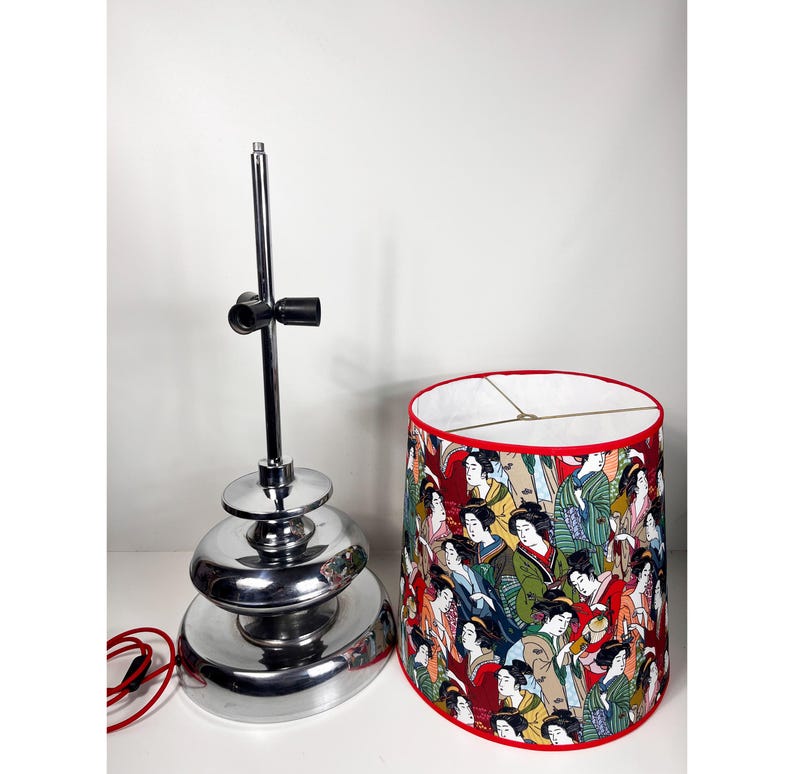Op de afbeelding: Een chromen tafellamp met een gelaagde basis en een rood-witte lampenkap met een ontwerp van vrouwen in traditionele Japanse kleding.