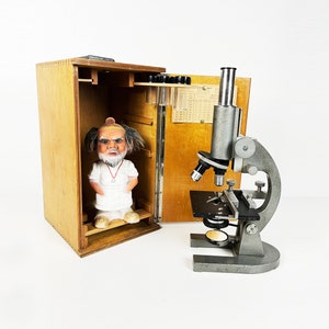 May include: A wooden box with a microscope and a small figurine of a man with a white beard and a white shirt. The box has a glass window and a small shelf inside. The microscope is silver and has a black base.