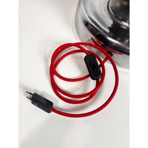 Op de afbeelding: Een rood elektrisch snoer met een zwarte in-line schakelaar en een zwarte stekker. Het snoer is opgerold en de stekker is zichtbaar.