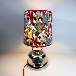 Op de afbeelding: Een zilveren tafellamp met een rood-witte lampenkap met een ontwerp van Japanse vrouwen. De lampenkap heeft een rode rand.