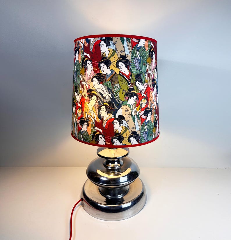 Op de afbeelding: Een zilveren tafellamp met een rood-witte lampenkap met een ontwerp van Japanse vrouwen. De lampenkap heeft een rode rand.