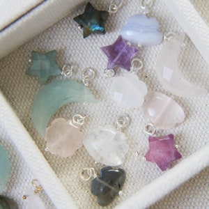 May include: A collection of gemstone charms in various shapes and colours, including stars, moons, and clouds. The charms are made of amethyst, labradorite, rose quartz, and other stones.