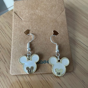 May include: A pair of silver dangle earrings with white and gold Mickey Mouse head charms. Each charm has a gold letter "M" in the center.