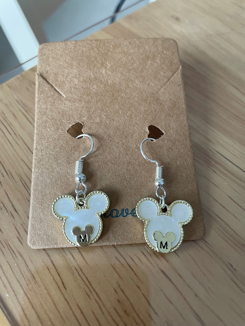 May include: A pair of silver dangle earrings with white and gold Mickey Mouse head charms. Each charm has a gold letter "M" in the center.