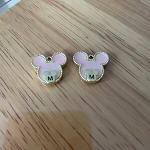 May include: Two pink and gold mouse-shaped charms with the letter "M" on each.