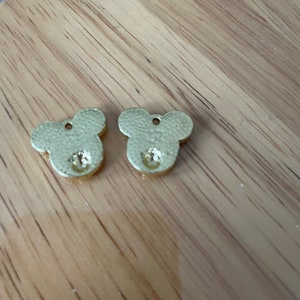 May include: Two gold-colored metal charms shaped like Mickey Mouse heads with a small, clear rhinestone in the center of each charm.