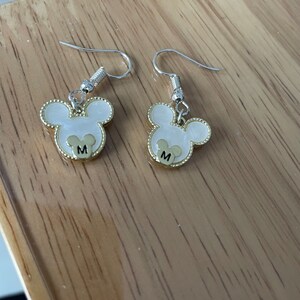 May include: A pair of silver dangle earrings with gold Mickey Mouse head charms. Each charm has a white center with a gold outline and the letter "M" in the center.