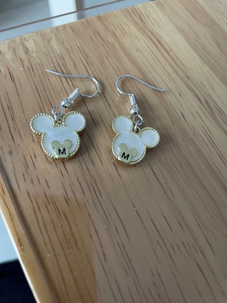 May include: A pair of silver dangle earrings with gold Mickey Mouse head charms. Each charm has a white center with a gold outline and the letter "M" in the center.