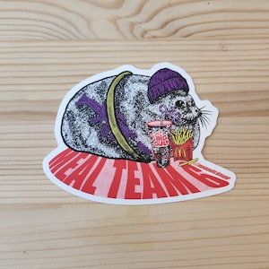 May include: A sticker featuring a cartoon seal wearing a purple beanie and holding a large cup of soda and a box of french fries. The seal is sitting on a red background with the text "Meal Team 6" in white letters.