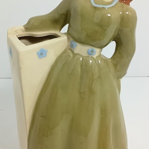 Puede incluir: Figura de cerámica de una mujer con un vestido largo verde, sosteniendo un jarrón crema. La mujer tiene el pelo rojo y una bufanda azul. Acentos de flores azules decoran el jarrón y la cintura de la mujer. La figura tiene un acabado brillante.