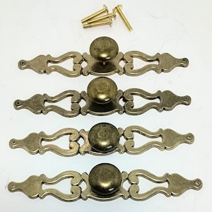 May include: Four brass-toned metal drawer pulls with a central round knob and an elongated, decorative backplate featuring open scrollwork. Each pull includes a matching brass-toned mounting screw positioned at the top of the arrangement.