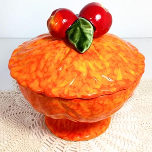 Può includere: Una zuppiera in ceramica arancione e gialla con coperchio e base a piedistallo. Il coperchio è sormontato da due ciliegie rosse e una foglia verde. La zuppiera ha una finitura lucida e poggia su una tovaglia di pizzo bianco.