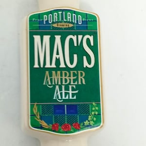 May include: A cream-colored beer tap handle with the text "PORTLAND BREWING" at the top. The main body features "MAC'S AMBER ALE" in white letters on a green background with decorative elements.