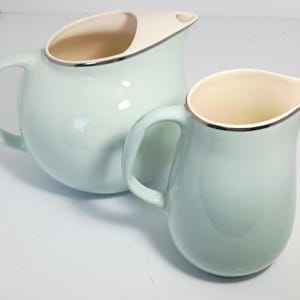 May include: Two ceramic jugs. The larger jug is light blue with a cream-coloured spout and interior. The rim is trimmed with silver. The smaller jug is light blue with a cream-coloured interior and silver trim.