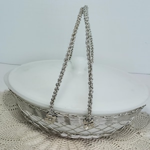 May include: An oval white glass casserole dish with a lid, encased in a silver-toned metal wire basket frame. The basket features a woven wire pattern, a tall hinged handle, and small metal flower accents where the handle attaches to the base.