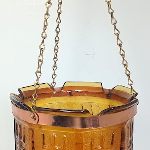 Op de afbeelding: Een hangende plantenbak van amberkleurig glas met een gestructureerd geometrisch patroon aan de buitenkant. De glazen houder wordt vastgehouden door een koperkleurige metalen band en hangt aan drie dunne goudkleurige metalen kettingen.