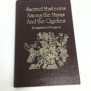Könnte beinhalten: Ein dunkelbraunes Hardcover-Buch mit goldener Schrift und einer goldenen Illustration einer Maya-Figur. Das Buch trägt den Titel "Sacred Mysteries Among the Mayas And the Quiches" von Augustus Le Plongeon.