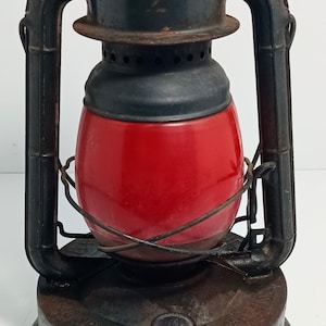 May include: Vintage lantern with a red glass globe and a dark metal frame. The lantern has a handle and a base with the word "SPECIAL" visible. The lantern is likely used for camping or as a decorative item.
