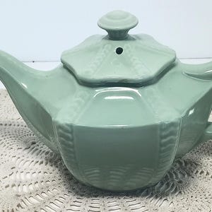Puede incluir: Una tetera de cerámica en color verde salvia claro con un cuerpo geométrico facetado y nervaduras verticales en relieve en los bordes. La tetera incluye una tapa a juego con un pomo central y un asa curva, con un acabado brillante y liso.