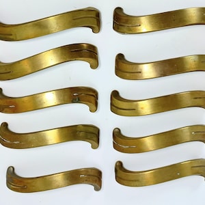 May include: Ten vintage brass drawer pulls with a curved, wave-like design. Each pull has a smooth, polished finish with subtle lines and a slight curve at each end. The brass has a warm, golden hue, suggesting age and use.