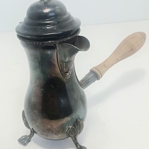 Puede incluir: Una cafetera de metal plateado con un acabado oscuro y deslustrado, que presenta una tapa abovedada, un pico curvo y un mango de madera clara. El recipiente se apoya sobre tres patas ornamentadas en forma de garra y tiene un cuerpo redondeado.