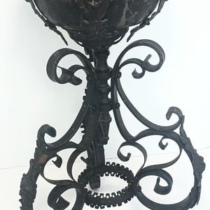 May include: A black metal pedestal planter featuring a rounded bowl with ornate relief details and lion head accents. The base is constructed from curved, scroll-like wrought iron legs that connect to a central circular ring near the bottom.