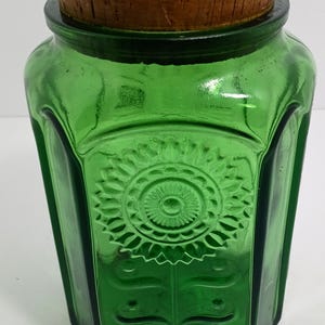 Puede incluir: Un tarro de vidrio verde con tapa de corcho de madera. El tarro tiene una forma geométrica con un diseño floral en relieve en la parte delantera. El tarro es probablemente vintage y adecuado para el almacenamiento.
