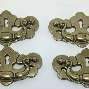 May include: Four brass-toned metal drawer pulls with a decorative backplate featuring a central keyhole cutout. Each pull has a curved, swinging handle attached to a scalloped, symmetrical plate with a muted, aged metallic finish.