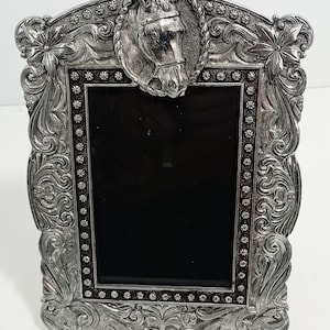 May include: Silver-colored picture frame with a horse head at the top. The frame has an ornate, detailed design with floral and scrollwork patterns. The frame's center is a black rectangle.