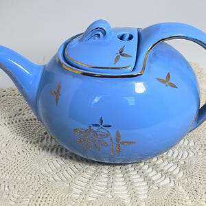 Puede incluir: Una tetera de cerámica azul claro con un cuerpo redondeado y un asa curvada. La tetera presenta detalles dorados en el borde, el pico y la tapa, y está decorada con diseños florales y de hojas doradas. La tetera está sobre un tapete de encaje blanco.