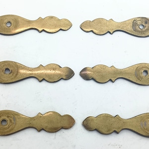 Könnte beinhalten: Sechs antike Schubladengriffe aus Messing mit einem dekorativen, gewellten Design. Jeder Griff hat ein zentrales Loch zur Montage. Das Messing hat eine warme, gealterte Patina, die auf einen Vintage- oder antiken Ursprung hindeutet. Ideal für die Möbelrestaurierung.