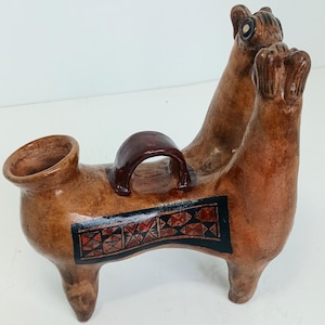 May include: A brown ceramic vessel shaped like a double-headed animal with a central handle. The body features a rectangular black panel with red and black geometric patterns. The piece has a smooth, polished finish and four short legs.