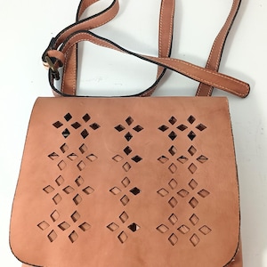May include: A light brown leather crossbody bag with a flap closure. The flap features a decorative cut-out pattern of diamonds and crosses. The bag has a long, adjustable strap and a small pocket on the front.