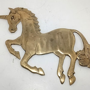 Puede incluir: Una decoración de pared plana de latón dorado con forma de unicornio en pose de salto. El unicornio presenta un cuerno largo y puntiagudo, una crin texturizada y una cola fluida. La superficie metálica muestra variaciones naturales de color.