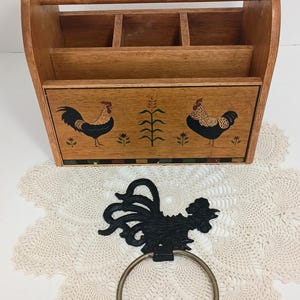 Könnte beinhalten: Holz-Organizer mit Griff und mehreren Fächern, mit Hahn-Illustrationen. Ein schwarzer Metall-Handtuchring in Form eines Hahns mit einem silbernen Ring ist ebenfalls vorhanden. Die Artikel befinden sich auf einer weißen Spitzendecke.