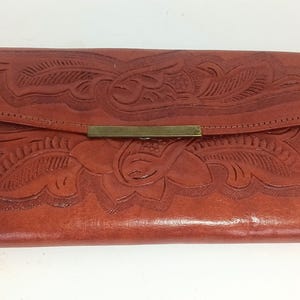 Könnte beinhalten: Eine braune Ledergeldbörse mit floralem Prägemuster. Die Geldbörse hat einen Klappverschluss mit einem messingfarbenen Metallverschluss. Das Leder scheint gealtert zu sein, mit sichtbarer Textur und einem satten, warmen Ton.